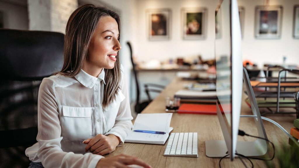 Woman working at a computer in an open-plan office, notebook in front of her.