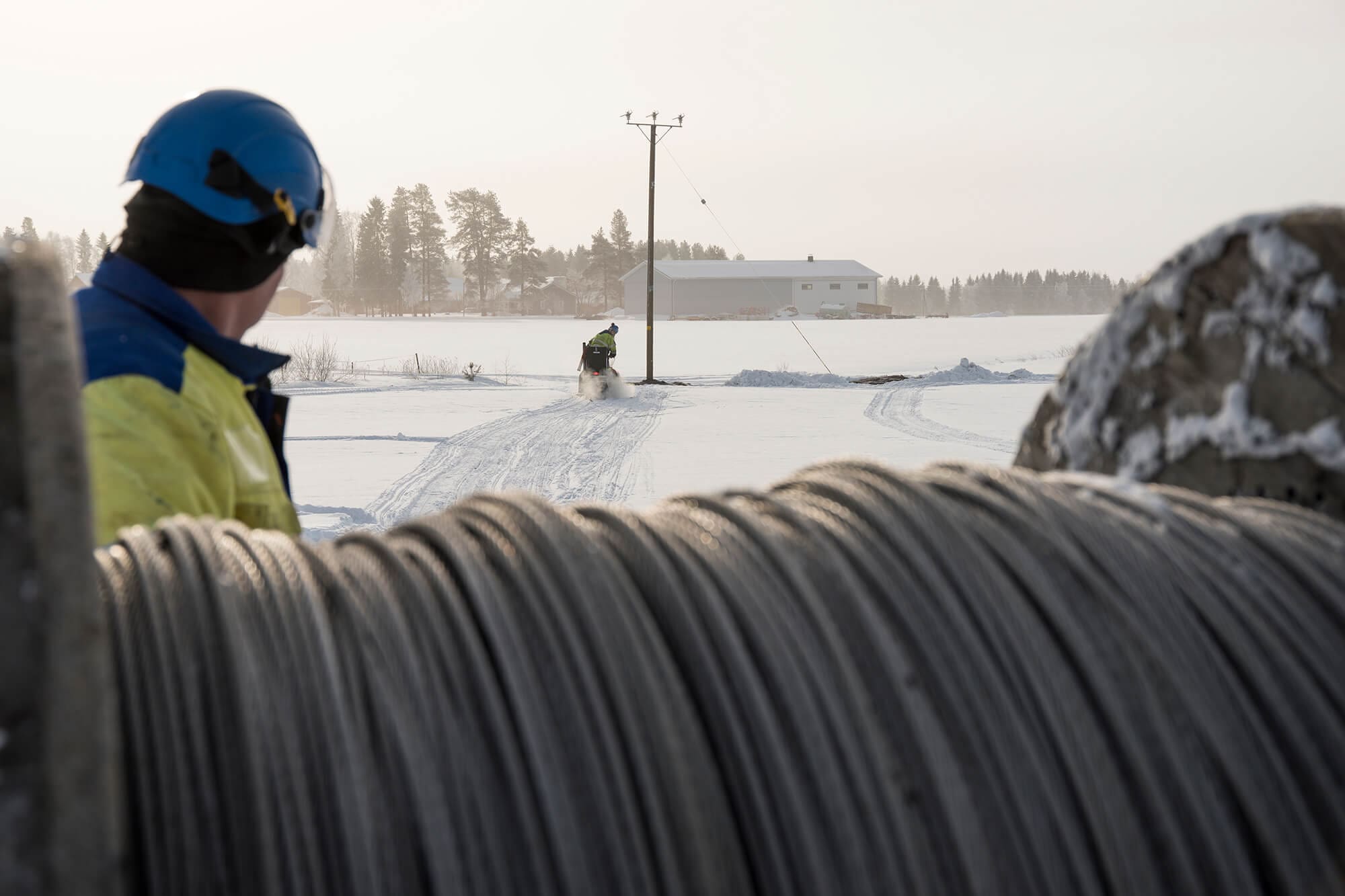 Työntekijät asentavat kaapelia lumisessa maastossa sähkölinjan varrella.